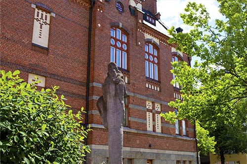 Lahden yhteiskoulun vanha koulurakennus Museo ja Olavi Lanun Yhteispuu-patsas, joita ympäröi kesän vihreys puiden ja pensaiden lehdistössä.