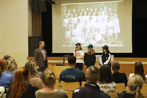 Koulun rehtori haastattelee tukioppilaita vanhempainillassa. Vanhempia yleisössä. Taustalla vanha mustavalkoinen valokuva koulun oppilaista.