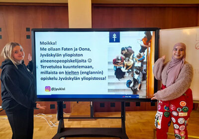 Kaksi tyttöä suuren näytön edessä. Toisella on yllä opiskelijahaalarit. Näytöllä lukee "Moikka! Me ollaan Faten ja Oona, Jyväskylän yliopiston aineenopeopiskelijoita. Tervetuloa kuuntelemaan, millaista on kielten (englannin) opiskelu Jyväskylän yliopistossa. Instagram-logo ja teksti: @jyukivi.