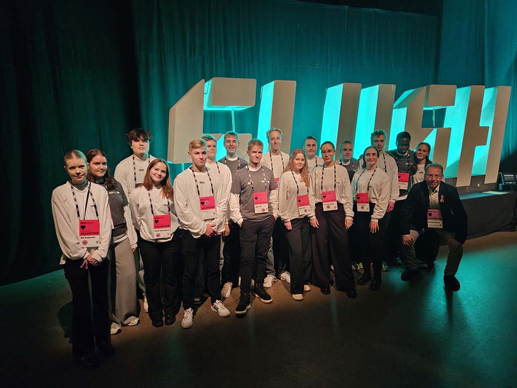 Opiskelijoita staff-kyltit kaulassa, taustalla Slush-valokyltti.