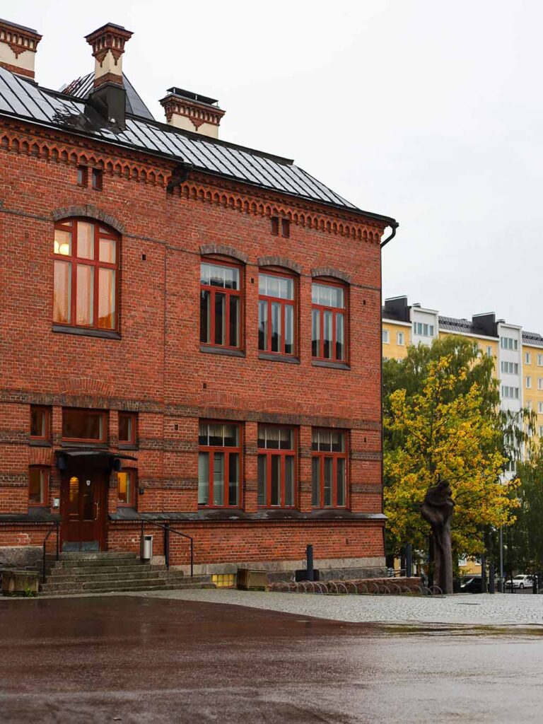 Lahden yhteiskoulun vanha koulurakennus Museo harmaana syyspäivänä kuvattuna. Taustalla on harmaa taivas ja puita, joissa ruskan väriset lehdet. Yhdessä ikkunassa loistaa valo, 