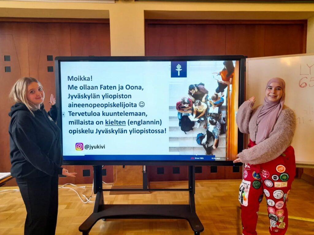 Kaksi tyttöä suuren näytön edessä. Toisella on yllä opiskelijahaalarit. Näytöllä lukee "Moikka! Me ollaan Faten ja Oona, Jyväskylän yliopiston aineenopeopiskelijoita. Tervetuloa kuuntelemaan, millaista on kielten (englannin) opiskelu Jyväskylän yliopistossa. Instagram-logo ja teksti: @jyukivi.
