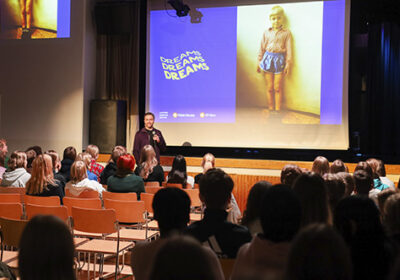 Koulun juhlasali, jossa paljon yleisöä istumassa penkeillä. Puhujana edessä keski-ikäinen valkoihoinen mies. Taustalla lapsuuden kuva pojasta heijastettuna valkokankaalle, jossa teksti Dreams, Dreams, Dreams (unelmia) aaltoilevalla fontilla.