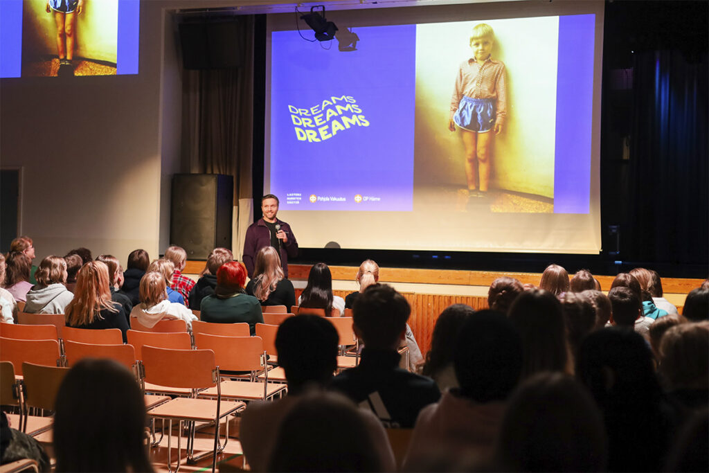 Koulun juhlasali, jossa paljon yleisöä istumassa penkeillä. Puhujana edessä keski-ikäinen valkoihoinen mies. Taustalla lapsuuden kuva pojasta heijastettuna valkokankaalle, jossa teksti Dreams, Dreams, Dreams (unelmia) aaltoilevalla fontilla.