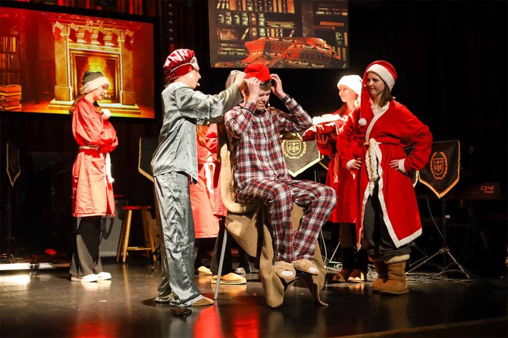 Joulujuhlanäytelmä koulun lavalla. Nuoria pyjamissa, tonttulakeissa ja yksi pukeutuneena joulupukiksi.