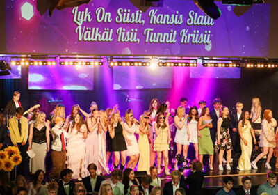 Lahden yhteiskoulun peruskoulun kevätjuhla 2026. Peruskoulun päättäviä oppilaita värivaloin valaistulla juhlasalin lavalla laulamassa. Seinälle on heijastettu laulun sanat: Lyk on siisti, Kansis staili. Välkät iisii, tunnit kriisii.